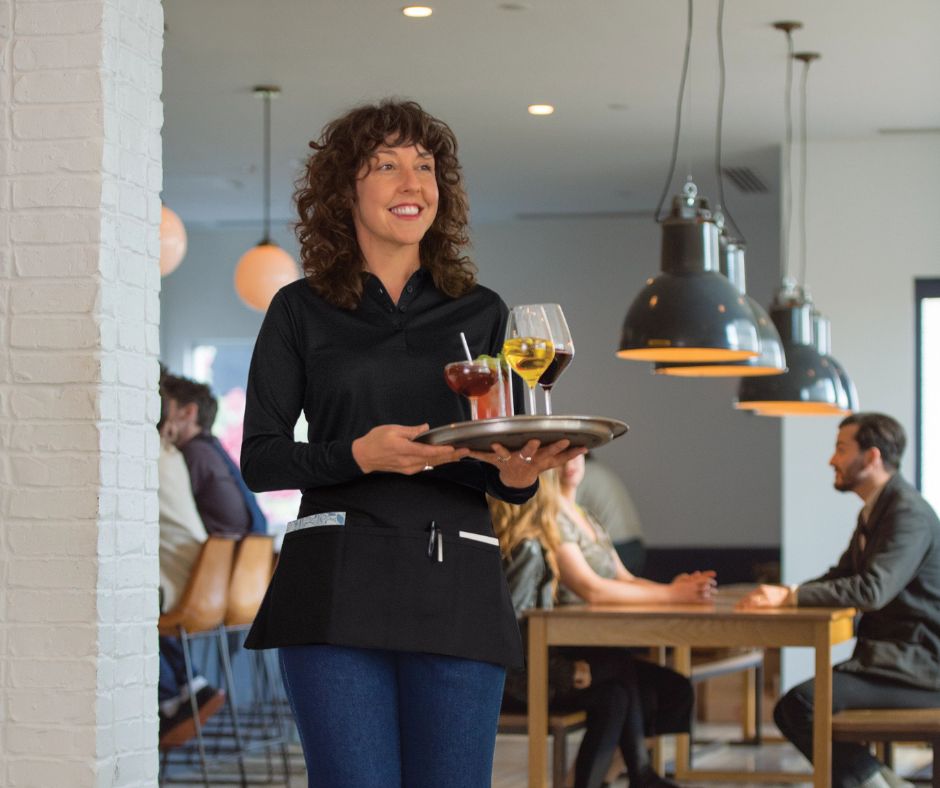 waitress carrying tray of drinks wearing a black button up shirt, black waist apron, and denim jeans